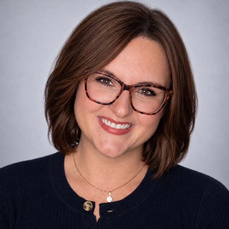 Portrait of a woman with brown shoulder-length hair wearing tortoiseshell glasses and a black top, smiling at the camera against a light gray background