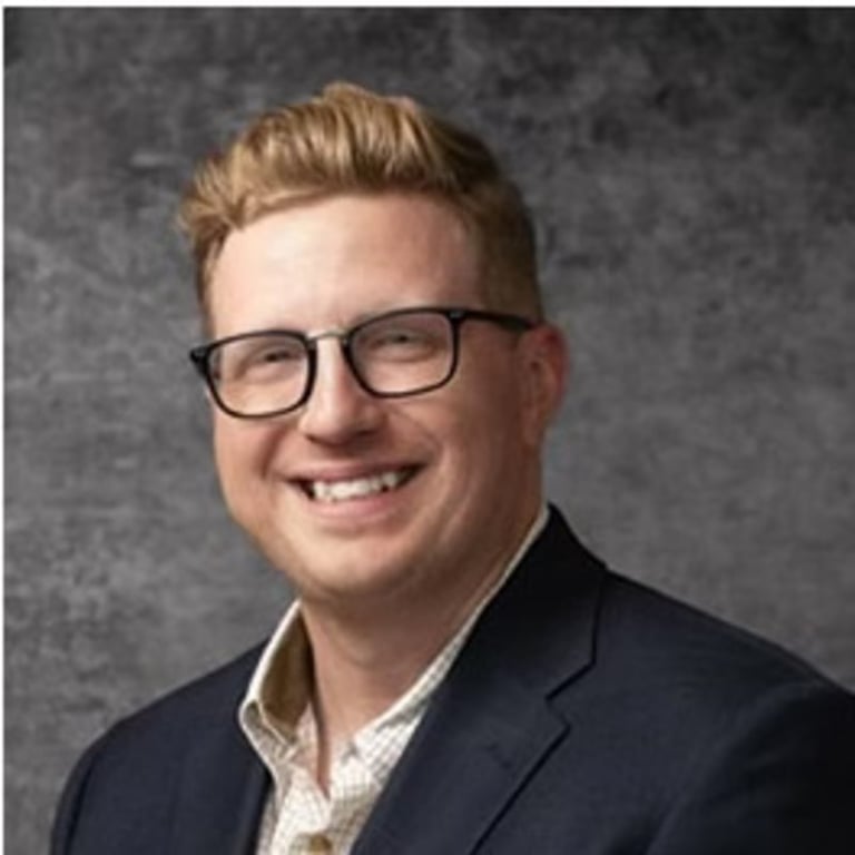 Professional headshot of a smiling man with blonde hair, glasses, and dark blazer against a grey background